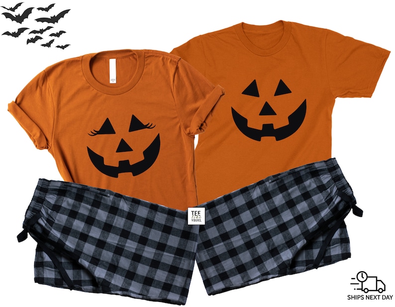 May include: A matching set of orange t-shirts with a black jack-o'-lantern face design. The t-shirt on the left has eyelashes on the jack-o'-lantern face. The set includes a pair of black and gray plaid pajama pants.