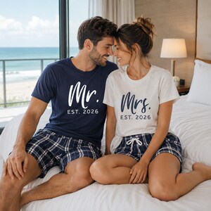Pu&ograve; includere: Una t-shirt blu navy con scritta bianca Mr. EST. 2026 e una t-shirt bianca con scritta blu navy Mrs. EST. 2026. Entrambe le maglie sono abbinate a pantaloncini da pigiama a quadri blu navy e bianchi con coulisse bianca in vita.