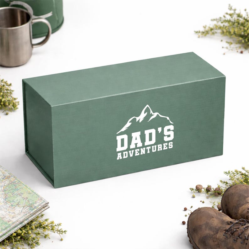May include: A rectangular, forest green gift box with the text "DAD'S ADVENTURES" in white, featuring a mountain graphic. The box is surrounded by outdoor-themed items, including a map, boots, and a metal mug.