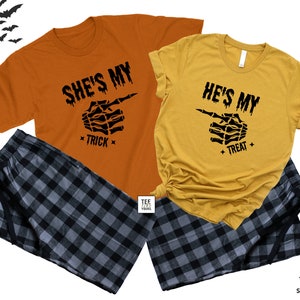 Op de afbeelding: Een bijpassende Halloween pyjamaset voor een stel, met oranje en gele shirts. De shirts hebben een skelethand die wijst met de tekst "She's My Trick" en "He's My Treat". De set bevat zwarte en grijze geruite pyjamabroeken.