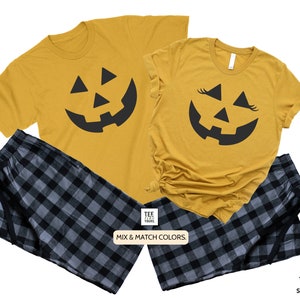 May include: A set of two mustard yellow t-shirts with a black jack-o'-lantern face graphic. One shirt has eyelashes on the jack-o'-lantern. The shirts are paired with black and gray plaid pajama pants.