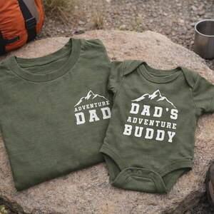 May include: Two olive green shirts with white text and mountain graphics. One is a t-shirt that says "ADVENTURE DAD", and the other is a baby onesie that says "DAD'S ADVENTURE BUDDY". The shirts are on a rock, with camping gear in the background.
