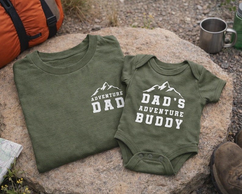 May include: Two olive green shirts with white text and mountain graphics. One is a t-shirt that says "ADVENTURE DAD", and the other is a baby onesie that says "DAD'S ADVENTURE BUDDY". The shirts are on a rock, with camping gear in the background.