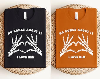 May include: Two t-shirts featuring a skeleton hand heart design with the text "No bones about it" and "I love her" on one shirt and "I love him" on the other.