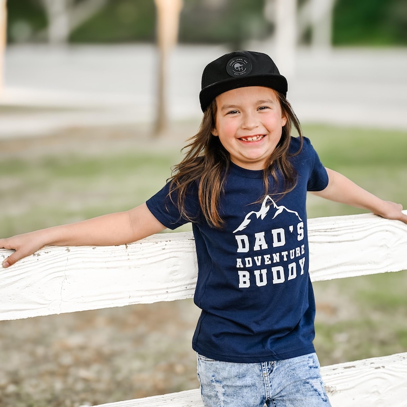May include: A child wearing a black cap and a navy blue t-shirt with the text "DAD'S ADVENTURE BUDDY" in white. The child is smiling and leaning on a white fence. The cap has a circular emblem.