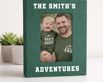 May include: A green photo album with the text "THE SMITH'S ADVENTURES" in white. The album holds a photo of a man and a baby. The baby wears a green onesie that says "DAD'S ADVENTURE BUDDY". The man wears a green shirt that says "ADVENTURE DAD".