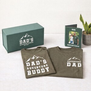 May include: A gift set featuring a green box and photo frame, along with two olive green shirts. The box and shirts have a mountain graphic and the text "DAD'S ADVENTURES". One shirt says "DAD'S ADVENTURE BUDDY". The photo frame says "OUR ADVENTURES".