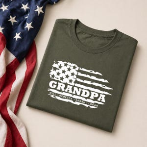 May include: An olive green t-shirt featuring a distressed American flag design with the word "GRANDPA" in bold white letters. Below the word are the names "WILL REBECCA SARAH LOGAN". An American flag is partially visible.