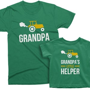 May include: Two green t-shirts with a cartoon tractor design. The larger shirt says "Grandpa" and the smaller shirt says "Grandpa's Little Helper".