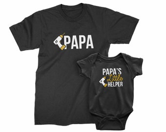May include: A black t-shirt with the word "PAPA" and a white drill icon. A black baby onesie with the text "PAPA'S Little HELPER" and a gold drill icon.