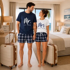 Pu&ograve; includere: Una t-shirt blu navy con scritta bianca Mr. EST. 2026 e una t-shirt bianca con scritta blu navy Mrs. EST. 2026. Entrambe sono abbinate a pantaloncini a quadri blu navy e bianchi con elastico in vita e coulisse regolabile.