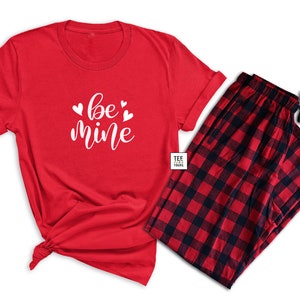 May include: A red pajama set featuring a short-sleeved t-shirt and plaid pajama pants. The t-shirt has the words "be mine" in white script with heart accents. The pajama pants are red and black checkered.