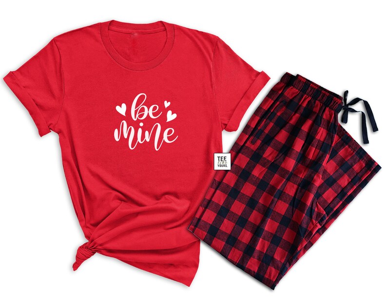 May include: A red pajama set featuring a short-sleeved t-shirt and plaid pajama pants. The t-shirt has the words "be mine" in white script with heart accents. The pajama pants are red and black checkered.