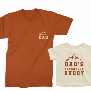 May include: Two matching t-shirts, one burnt orange and one off-white. The burnt orange shirt has "ADVENTURE DAD" printed on the chest. The off-white shirt has "DAD'S ADVENTURE BUDDY" printed on the front, both with a mountain graphic.