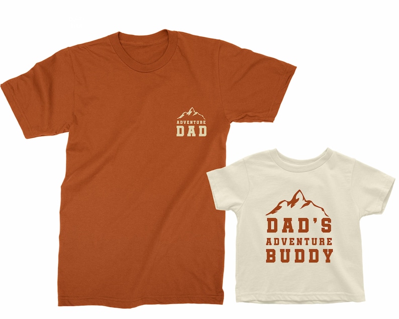 May include: Two matching t-shirts, one burnt orange and one off-white. The burnt orange shirt has "ADVENTURE DAD" printed on the chest. The off-white shirt has "DAD'S ADVENTURE BUDDY" printed on the front, both with a mountain graphic.