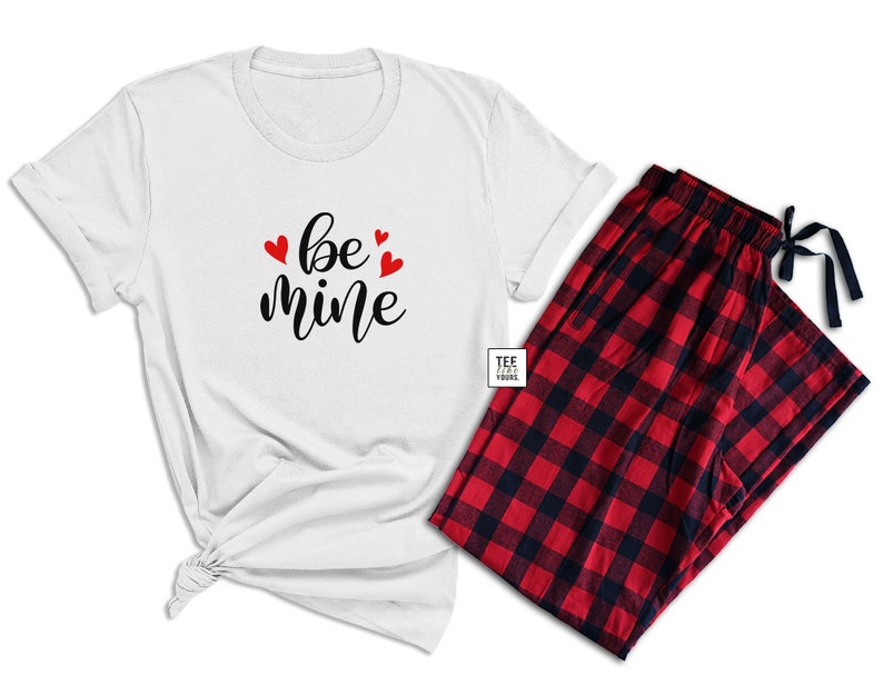 May include: A white t-shirt with the words "be mine" in black script and red heart accents, paired with red and black plaid pajama pants. The pajama pants have a drawstring closure.