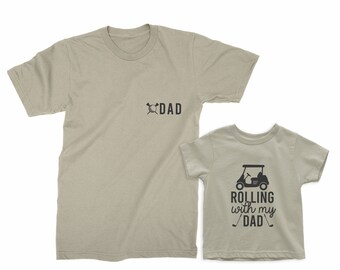 May include: A set of two tan t-shirts with a golf cart design. The larger shirt says "DAD" with a golf club and ball. The smaller shirt says "ROLLING with my DAD" with a golf cart and golf clubs.