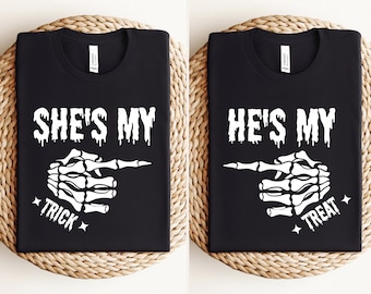 May include: Two black t-shirts with white text and a skeleton hand graphic. The left shirt says "She's My Trick" and the right shirt says "He's My Treat".