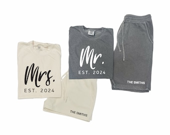 May include: Two matching t-shirts and shorts sets for a couple. The t-shirts are white and gray with black text that says "Mrs. EST. 2024" and "Mr. EST. 2024". The shorts are white and gray with black text that says "THE SMITHS".