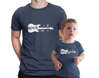 Könnte beinhalten: Passende marineblaue T-Shirts für Vater und Kind. Das Shirt des Vaters trägt den Schriftzug "Rock And" mit einer weißen Silhouette einer E-Gitarre. Das Shirt des Kindes trägt den Schriftzug "Roll Baby" mit einer weißen Silhouette einer E-Gitarre.