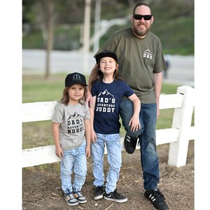 May include: A family poses outdoors, wearing matching shirts and hats. The shirts read "DAD'S ADVENTURE BUDDY" with a mountain graphic. The dad wears a green shirt with "ADVENTURE DAD" and sunglasses. The children wear jeans and hats.