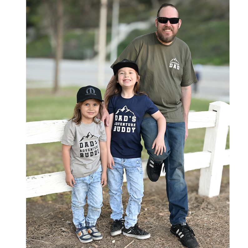 May include: A family poses outdoors, wearing matching shirts and hats. The shirts read "DAD'S ADVENTURE BUDDY" with a mountain graphic. The dad wears a green shirt with "ADVENTURE DAD" and sunglasses. The children wear jeans and hats.