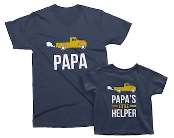 May include: Two navy blue t-shirts with a yellow truck graphic. The larger shirt says "PAPA" and the smaller shirt says "PAPA'S LITTLE HELPER".