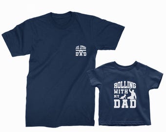 May include: Two navy blue t-shirts. One adult shirt has "Jiu Jitsu Dad" printed in white. The other is a child's shirt with "Rolling with my Dad" and a graphic of a person and child in white.