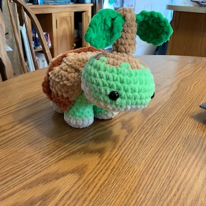 May include: A handmade crochet turtle plushie in shades of green, brown, and tan. The turtle has a textured shell, a green head, and small black eyes. The plushie is sitting on a wooden table.