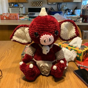 May include: A crocheted plush toy with a red body, white ears, and a pink nose. The toy has a horn on its head and is sitting on a wooden surface.