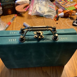 Puede incluir: Un diario encuadernado en cuero color turquesa con un cierre de latón y dos asas. El diario tiene costuras decorativas a lo largo de los bordes.