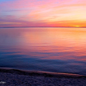 May include: A vibrant sunset over a calm body of water, with the sky transitioning from deep blue to pink and orange. The water reflects the colours of the sky, creating a serene and colourful scene.