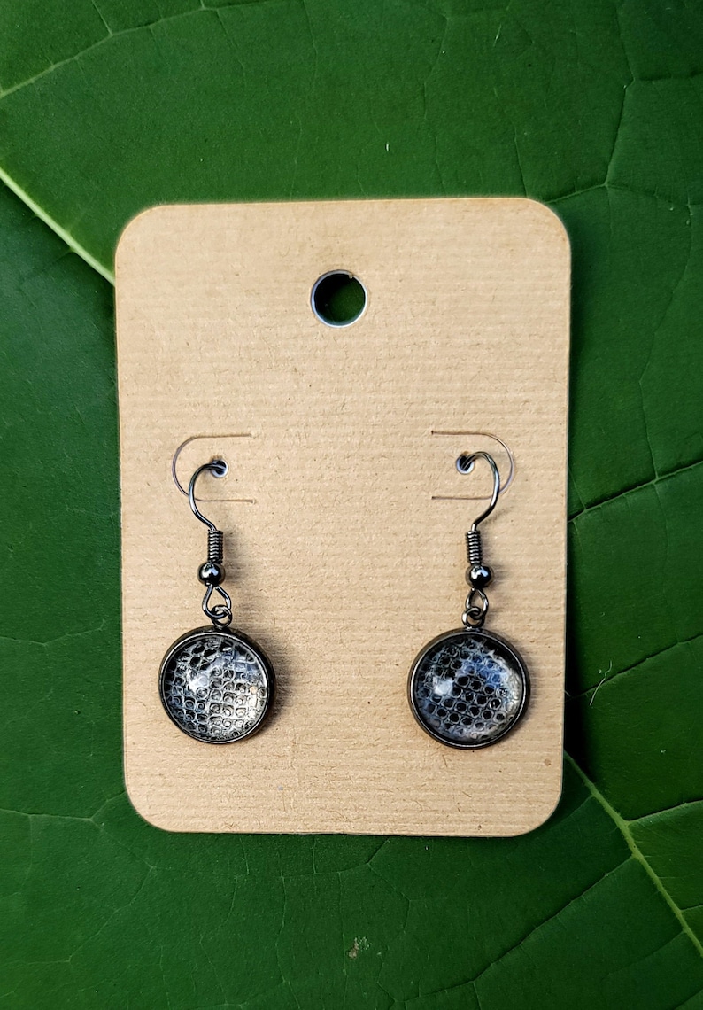 Puede incluir: Un par de pendientes colgantes de plata con un colgante circular estampado en blanco y negro.