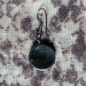 Puede incluir: Un pendiente de plata con un dije redondo de metal negro. El pendiente tiene un cierre simple de gancho.