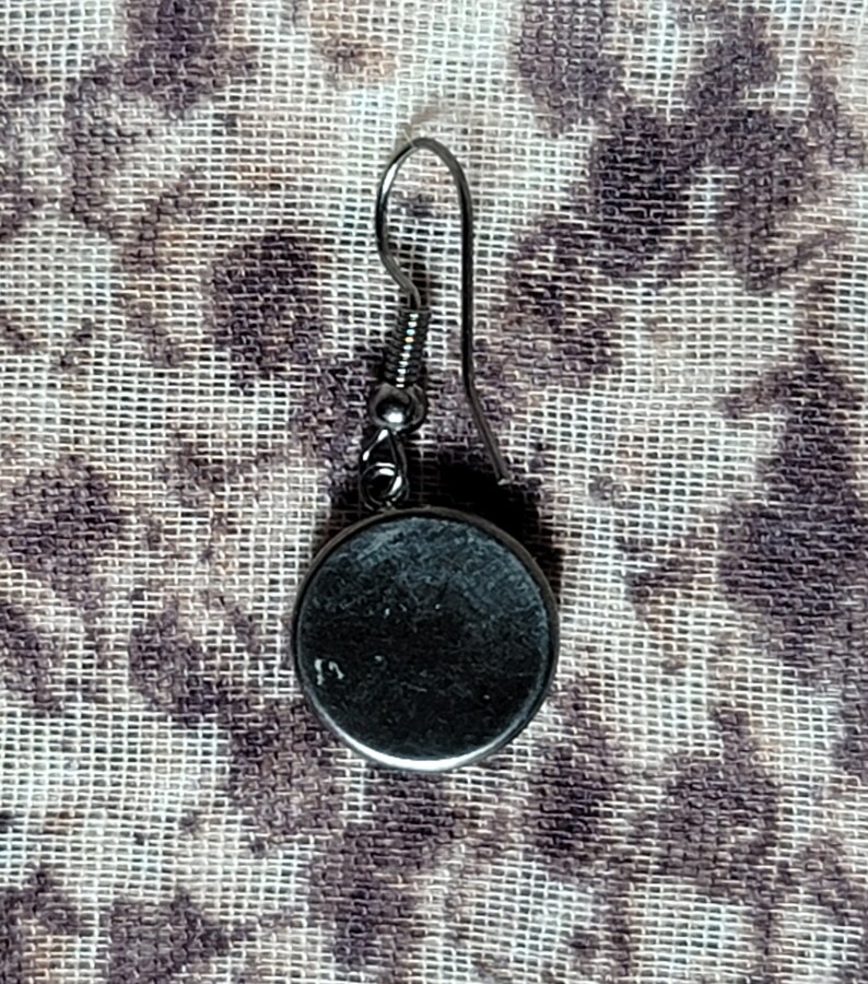 Puede incluir: Un pendiente de plata con un dije redondo de metal negro. El pendiente tiene un cierre simple de gancho.