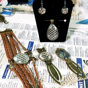 Puede incluir: Un conjunto de joyas con un patr&oacute;n de piel de serpiente. El conjunto incluye un collar con un colgante, un par de pendientes y una pulsera. El colgante tiene forma de l&aacute;grima con un engaste plateado. Los pendientes son de plata con un patr&oacute;n de piel de serpiente. La pulsera es de cuero marr&oacute;n con un dije de piel de serpiente.