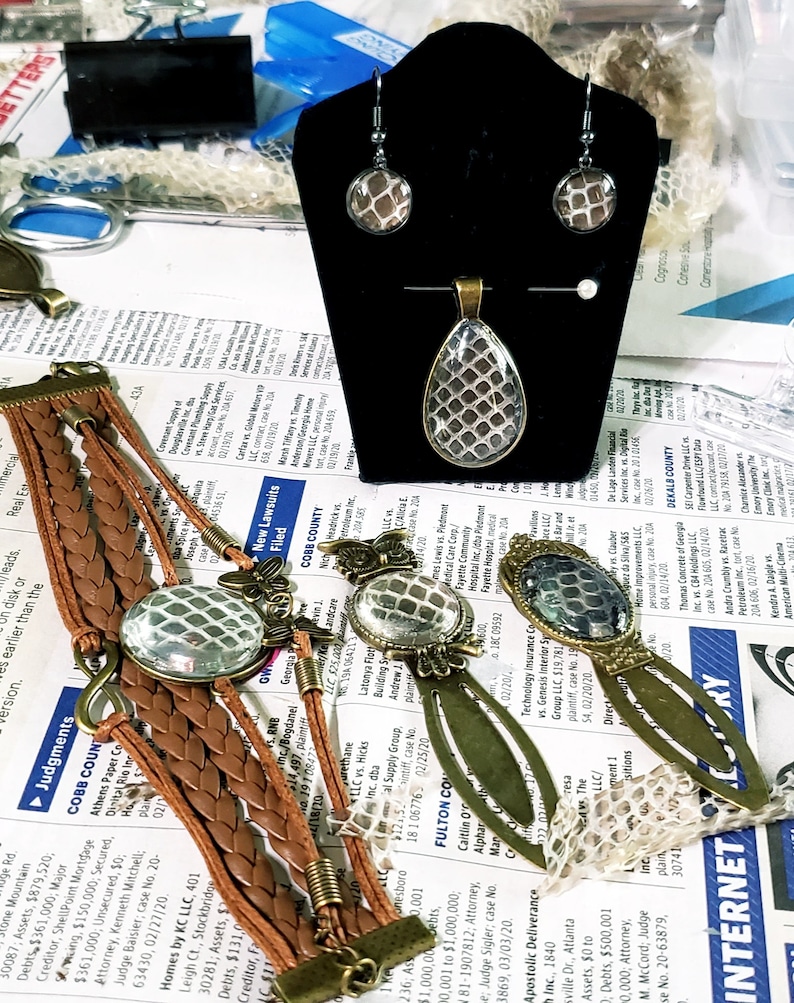 Puede incluir: Un conjunto de joyas con un patr&oacute;n de piel de serpiente. El conjunto incluye un collar con un colgante, un par de pendientes y una pulsera. El colgante tiene forma de l&aacute;grima con un engaste plateado. Los pendientes son de plata con un patr&oacute;n de piel de serpiente. La pulsera es de cuero marr&oacute;n con un dije de piel de serpiente.