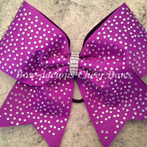 May include: Purple cheer bow with silver rhinestones and a black elastic band. The bow is decorated with the text "Bow-da-cious Cheer Bows".