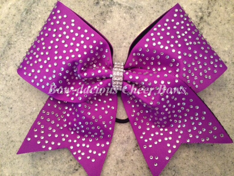 May include: Purple cheer bow with silver rhinestones and a black elastic band. The bow is decorated with the text "Bow-da-cious Cheer Bows".