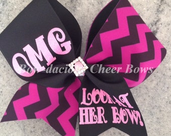 May include: A large black and pink cheer bow with a chevron pattern and the text "OMG" and "LOOK AT HER BOW!". The bow features a rhinestone embellishment in the center.