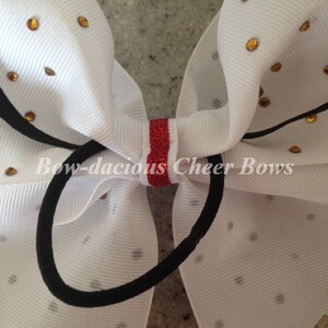 May include: White cheer bow with gold glitter dots and a red glitter stripe in the center. The bow is tied with a black elastic band. The text "Bow-dacious Cheer Bows" is printed on the bow.