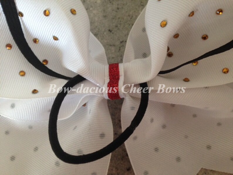 May include: White cheer bow with gold glitter dots and a red glitter stripe in the center. The bow is tied with a black elastic band. The text "Bow-dacious Cheer Bows" is printed on the bow.