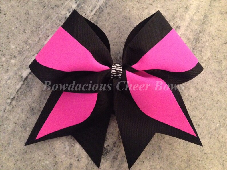 May include: A large black and hot pink cheer bow with a silver rhinestone in the center. The bow is made of two layers of ribbon and has a pointed shape.