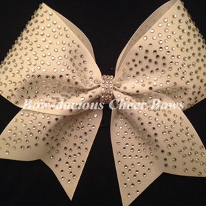 May include: A large ivory cheer bow with a center rhinestone and a pattern of rhinestones throughout the bow. "Bow-da-cious Cheer Bows" is written on the bow.