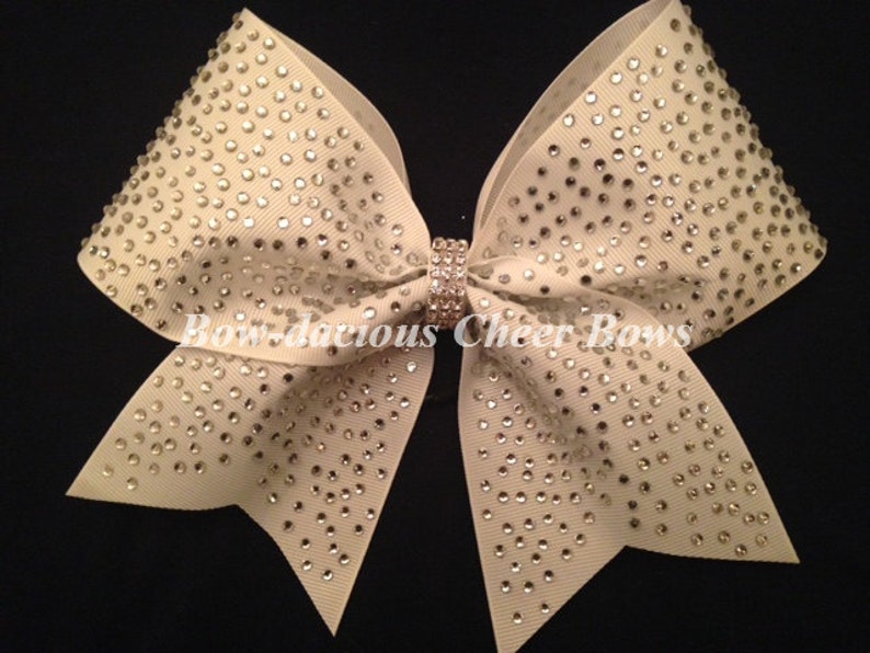 May include: A large ivory cheer bow with a center rhinestone and a pattern of rhinestones throughout the bow. "Bow-da-cious Cheer Bows" is written on the bow.