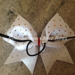 May include: White cheer bow with gold glitter and rhinestones. The bow is tied with a black elastic band and has a red and white stripe in the center.