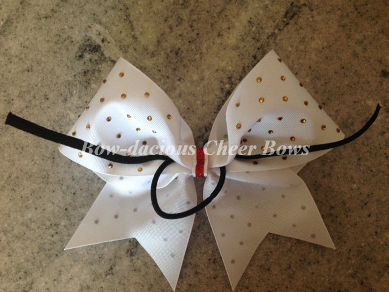 May include: White cheer bow with gold glitter and rhinestones. The bow is tied with a black elastic band and has a red and white stripe in the center.