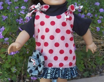 May include: A doll wearing a black top with a red and white polka dot dress. The dress has a black and white patterned ruffle at the hem. The doll has a matching bow in her hair and black patent leather shoes.