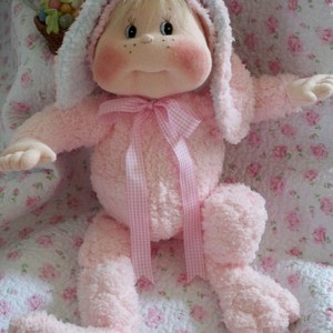 May include: A plush pink bunny doll with a hood and bow. The doll has a round face with rosy cheeks, freckles, and a small mouth. It is wearing a pink outfit with a gingham ribbon and has long, floppy ears.