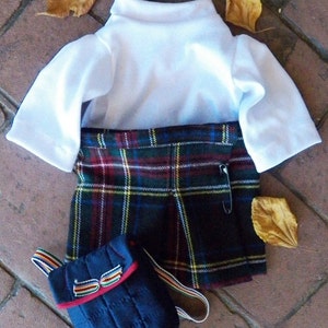 May include: A doll-sized school uniform set featuring a white short-sleeved shirt, a plaid skirt with a safety pin, and a small navy blue backpack with colorful straps. The skirt has a green, red, and yellow plaid pattern.